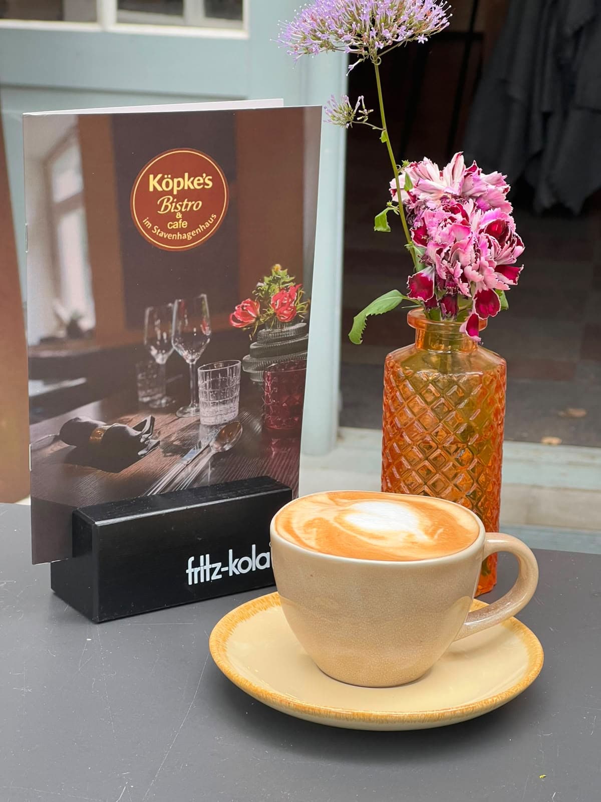 Cappuccino mit Milchschaum auf einem Tisch, daneben eine orangefarbene Glasvase mit rosa Blüten und die Karte mit dem Köpke’s-Logo